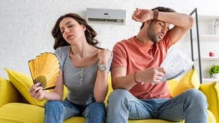 Mann und Frau sitzen schwitzend auf der Couch und fächern sich Luft zu. Symbolbild Test rotorloser Ventilatoren.