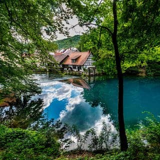 Der Blautopf bei Ulm ist ein schönes Ausflugsziel in Baden-Württemberg. Der blaue See liegt im Wald vor einem Bauernhaus mit Fachwerk.