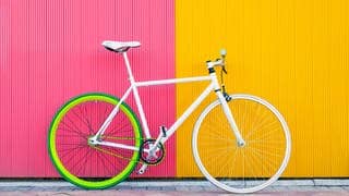 Ein weißes Fahrrad steht vor einer gelb-pinken Wand. 