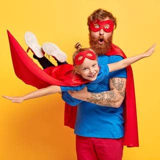 Mann, verkleidet als Superheld, hält sein Kind in die Luft, das ebenfalls als Heldin verkleidet ist. Symbolbild für Helden des Alltags.