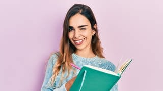 Junge Frau hält Buch und Stift in der Hand und zwinkert. Symbolbild für unübersetzbare deutsche Wörter, die es in anderen Sprachen nicht gibt.