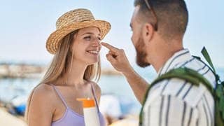 Mann und Frau stehen am Strand. Er cremt ihr die Nase mit Sonnencreme ein. Symbolbild für Stiftung Warentest zu Sonnencremes.