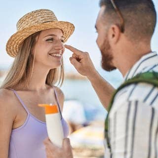 Mann und Frau stehen am Strand. Er cremt ihr die Nase mit Sonnencreme ein. Symbolbild für Stiftung Warentest zu Sonnencremes.