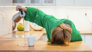 Junge Frau steht in der Küche, mit dem Kopf auf dem Tisch. Sie schüttet Kaffee aus der Maschine NEBEN die Kaffeetasse. Symbolbild für wenig Schlaf und große Müdigkeit.