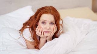 Junge Frau liegt im Bett. Sie hält sich das Gesicht müde mit den Händen. Symbolbild für wenig Schlaf und Müdigkeit – wie kann ich jetzt den Arbeitstag überstehen?