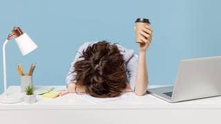 Junge Frau liegt müde mit dem Kopf auf dem Schreibtisch, eine Hand mit Kaffeebecher streckt sie in die Hand. Symbolbild für wenig Schlaf und Müdigkeit im Arbeitstag.