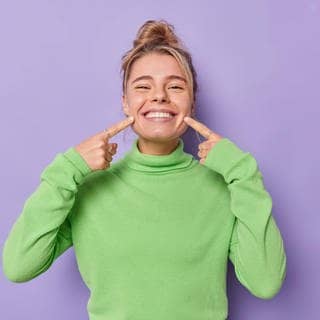 Eine Frau zeigt ihr Lachen und deutet mit den Fingern auf den Mund. Symbolbild für die aktuellen Testergebnisse 2024 der Stiftung Warentest zu Zahnzusatzversicherungen.