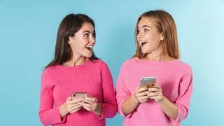 Zwei junge Frauen halten ein Smartphone in der Hand und lachen. Symbolbild für kuriose Redewendungen aus anderen Sprachen der Welt.