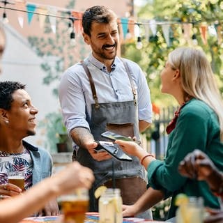 Gruppe von jungen Menschen in einem Restaurant, sieht nach Urlaub aus. Eine Frau bezahlt und gibt dem Kellner Trinkgeld.