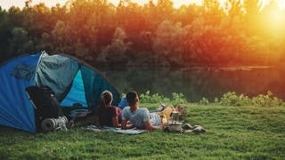 Junge Frau und junger Mann liegen neben einem Zelt im Gras und schauen auf den Sonnenuntergang. Symbolbild: Tipps fürs Camping für Anfänger mit Packliste und Checkliste.