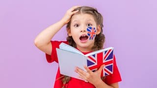 Kleines Mädchen mit Zöpfchen guckt erstaunt und fasst sich mit der Hand an den Kopf. Sie hält ein Buch mit englischer Flagge in der Hand, auf der Wange ist sie bemalt. Symbolbild für denglische Wörter und deren ganz andere Bedeutung auf Englisch.