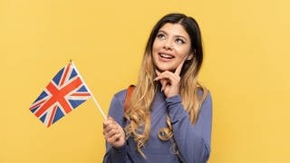 Junge Frau schaut fragend, in der Hand hält sie eine England-Flagge. Symbolbild für denglische Wörter, die auf Englisch eine ganz andere Bedeutung haben.