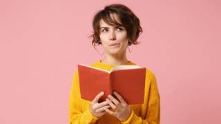 Junge Frau hält ein Buch in der Hand und schaut denkend in die Luft. Symbolbild für: Duden-Quiz – welche Wörter sind neu dazugekommen, welche wurden gestrichen?