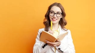 Junge Frau hält denkend einen Stift ans Kinn und einen Schreib-Block in der Hand. Symbolbild für einen Artikel zum Thema Komma und Sätze, deren Bedeutung sich verändert, wenn man die Kommata verschiebt.