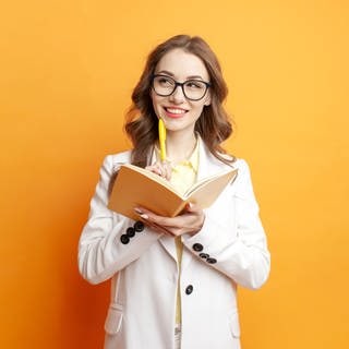 Junge Frau hält denkend einen Stift ans Kinn und einen Schreib-Block in der Hand. Symbolbild für einen Artikel zum Thema Komma und Sätze, deren Bedeutung sich verändert, wenn man die Kommata verschiebt.