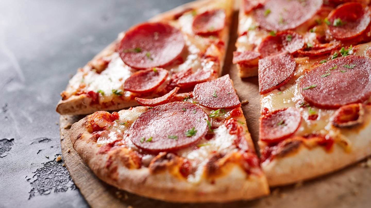 Eine Salami-Tiefkühlpizza auf einem Holzbrett und schwarzem, steinernen Untergrund. Stiftung Warentest hat 21 Tiefkühlpizzen auf Geschmack, ernährungsphysiologische Merkmale und weiteren Kriterien getestet.