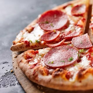 Eine Salami-Tiefkühlpizza auf einem Holzbrett und schwarzem, steinernen Untergrund. Stiftung Warentest hat 21 Tiefkühlpizzen auf Geschmack, ernährungsphysiologische Merkmale und weiteren Kriterien getestet.