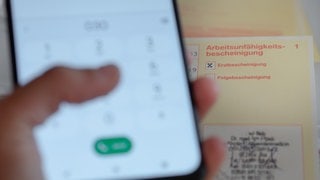 Krankmeldung beziehungsweise Arbeitsunfähigkeitsbescheinigung für den Arbeitgeber. Im Vordergrund hält jemand ein Handy in der Hand.