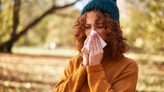 Uma mulher usando um chapéu de tricô azul e um suéter amarelo espirra em seu lenço porque está gripada ou resfriada e não conseguiu se proteger contra isso