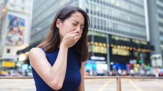 Frau vor Großstadtkulisse hat Übelkeit, eventuell durch Migräne oder Kopfschmerzen bedingt, und hält sich die Hand vor den Mund.