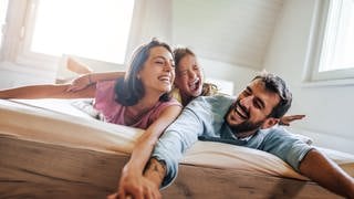 Eine junge Familie liegt auf einer Matratze in ihrem Bett und tollt umher. Vater, Mutter und das Kind freuen sich und haben ein Lächeln im Gesicht.