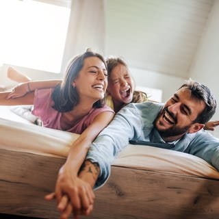 Eine junge Familie liegt auf einer Matratze in ihrem Bett und tollt umher. Vater, Mutter und das Kind freuen sich und haben ein Lächeln im Gesicht.