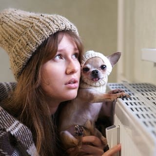 Eine junge Frau sitzt in eine Decke gehüllt und mit Mütze auf dem Kopf an der Heizung und scheint zu frieren. Auch ihr kleiner Hund hat eine Wintermütze auf. Symbolbild für die Frage, ab wann der Vermieter dem Mieter die Heizung anschalten muss. Wie sind die Regelungen in der Heizperiode?