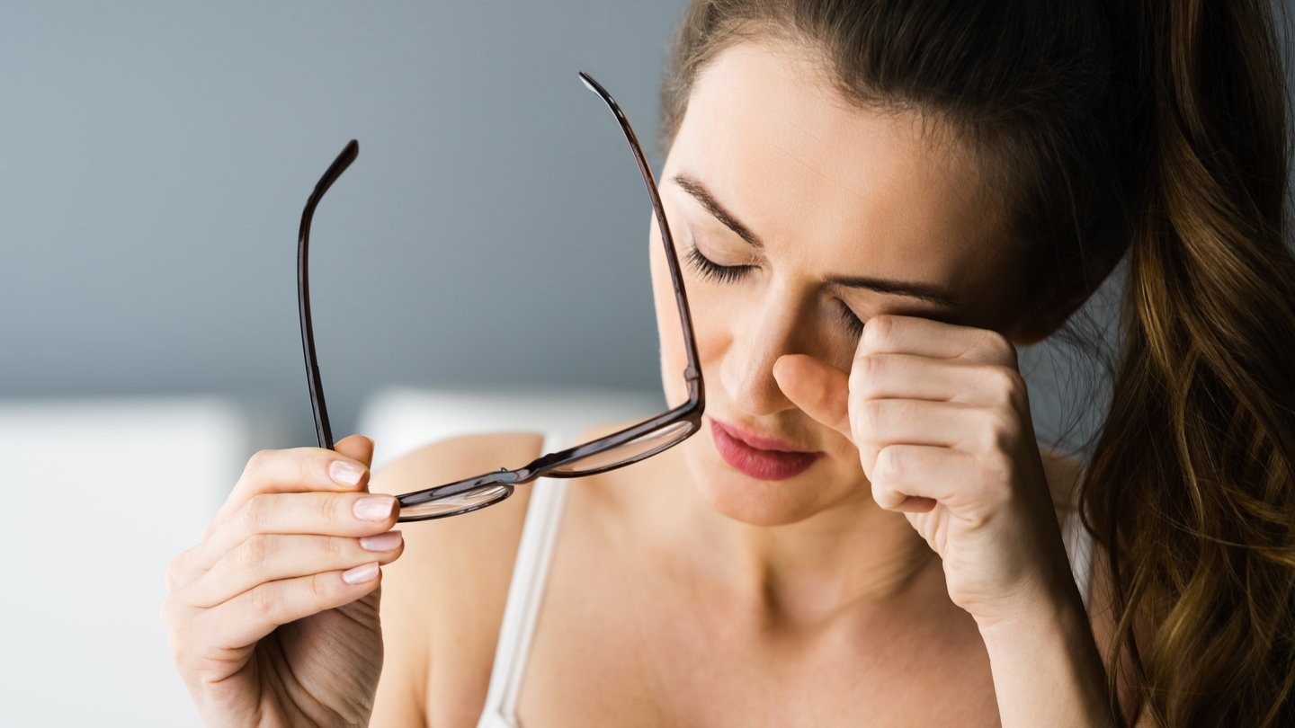 Eine junge Frau sieht sehr müde aus, hat sich die Brille heruntergezogen und reibt sich die Augen. Symbolbild für Long Covid bzw. Post Covid. Bei der Krankheit zählt chronische Müdigkeit (Chronisches Fatigue Syndrom) zum einem der häufigtsten Symptome.