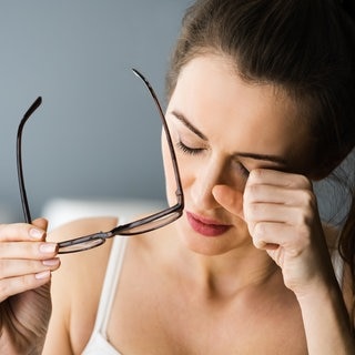 Eine junge Frau sieht sehr müde aus, hat sich die Brille heruntergezogen und reibt sich die Augen. Symbolbild für Long Covid bzw. Post Covid. Bei der Krankheit zählt chronische Müdigkeit (Chronisches Fatigue Syndrom) zum einem der häufigtsten Symptome.