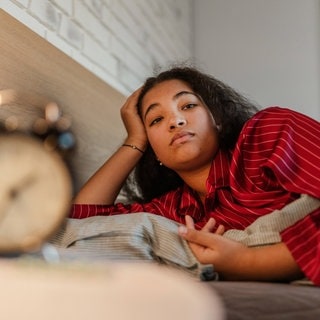 Eine Frau in rotem Pyjama liegt im Bett und kann nicht schlafen. Sie schaut genervt zum den Wecker, der halb 3 Uhr nachts anzeigt. Symbolbild für die Frage, ob wir bei Vollmond wirklich schlechter schlafen. Wir haben dazu mit Schlafforschern gesprochen und auch nachgefragt, ob die Schlafprobleme bei einem Supermond noch schlimmer werden.