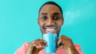 Ein junger Mann mit einer blauen Tasse mit einem Heißgetränk darin steht vor einem türkisen Hintergrund und lächelt. Symbolbild für die Testergebnisse bei Stiftung Warentest. Dort wurden 15 Filterkaffees getestet.