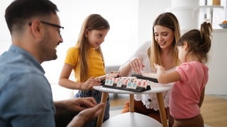 Mama und Papa spielen zusammen mit den Kindern ein Brettspiel (Rummikub) und lachen. Symbolfoto für das Thema, ob man als Erwachsene bzw. Eltern Kindern gewinnen oder verlieren lassen sollte und wie Kinder die Konzepte von gewinnen und verlieren am besten lernen können.