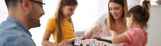 Mama und Papa spielen zusammen mit den Kindern ein Brettspiel (Rummikub) und lachen. Symbolfoto für das Thema, ob man als Erwachsene bzw. Eltern Kindern gewinnen oder verlieren lassen sollte und wie Kinder die Konzepte von gewinnen und verlieren am besten lernen können.