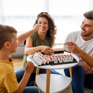 Mama und Papa spielen zusammen mit den Kindern ein Brettspiel (Rummikub) und lachen. Symbolfoto für das Thema, ob man als Erwachsene bzw. Eltern Kindern gewinnen oder verlieren lassen sollte und wie Kinder die Konzepte von gewinnen und verlieren am besten lernen können.