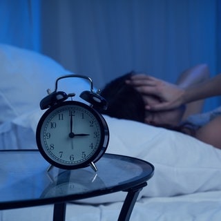 Junge Frau liegt nachts wach im Bett, auf ihrer Uhr steht 3 Uhr nachts. Symbolbild für eine Erklärung der Hintergründe zur Wolfsstunde oder Hexenstunde, die vielen beim Schlaf Probleme macht.