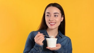 Junge Frau löffelt fragend einen Joghurt aus. Symbolbild fürs Grammatik-Quiz: der, die oder das – welcher Artikel ist richtig im Deutschen?