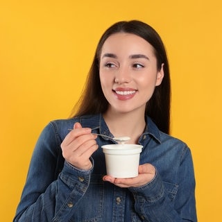 Junge Frau löffelt fragend einen Joghurt aus. Symbolbild fürs Grammatik-Quiz: der, die oder das – welcher Artikel ist richtig im Deutschen?