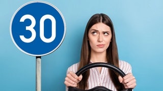 Junge Frau sitzt denkend hinter dem Lenkrad und schaut auf ein blaues Verkehrsschild mit weißer Schrift. Symbolbild für unbekannte Verkehrsschilder in Deutschland und ihre Bedeutung für Autofahrer.