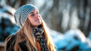 Junge Frau mit Mütze in Winterlandschaft wird von der Sonne beschienen. Symbolbild für Vitamin-D-Mangel, sie Symptome und was man dagegen tun kann.