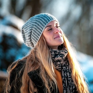 Junge Frau mit Mütze in Winterlandschaft wird von der Sonne beschienen. Symbolbild für Vitamin-D-Mangel, sie Symptome und was man dagegen tun kann.