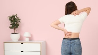 Eine junge Frau steht mit dem Rücken zur Kamera gewand in einem Zimmer mit einer kleinen Kommode und fasst sich an Nacken und unteren Rücken. Symbolbild für Rückenschmerzen unten denen ganz viele Menschen leiden. Doch es gibt einfache Übungen, die bei Rückenschmerzen gut helfen können.