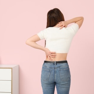 Eine junge Frau steht mit dem Rücken zur Kamera gewand in einem Zimmer mit einer kleinen Kommode und fasst sich an Nacken und unteren Rücken. Symbolbild für Rückenschmerzen unten denen ganz viele Menschen leiden. Doch es gibt einfache Übungen, die bei Rückenschmerzen gut helfen können.