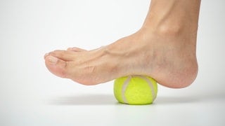 Ein Tennisball unter der Fußsohle eines nackten Fußes. Dabei geht es um das Lockern von verspannten Faszien im Fuß. Auch dies kann gegen Rückenschmerzen helfen.