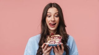 Junge Frau schaut hungrig auf ein Stück Torte. Symbolbild zum Mythos Dessert-Magen: Warum geht ein Nachtisch oder Süßes immer noch rein, obwohl man satt ist?