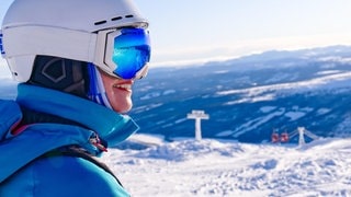 Eine Frau steht mit Skiausrüstung auf dem Berg und freut sich, weil im Skigebiet das Wetter passt