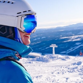 Eine Frau steht mit Skiausrüstung auf dem Berg und freut sich, weil im Skigebiet das Wetter passt