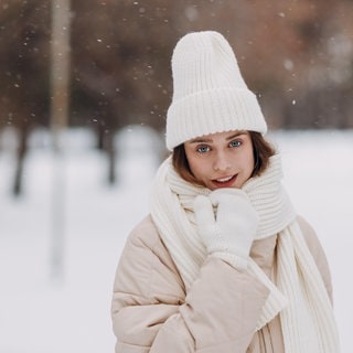 Eine Frau steht umgeben von Schnee in warmer Kleidung in der Natur und freut sich, dass in den Skigebieten das Wetter passt