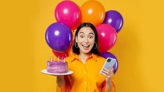 Junge Frau mit Handy und Geburtstagstorte in der Hand und Luftballons im Hintergrund. Symbolbild für lustige Whatsapp-Sprüche und Grüße zum Geburtstag.