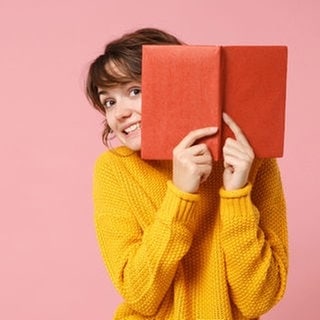 Eine junge Frau schaut hinter einem Buch hervor und freut sich über die SWR3 Buchtipps 