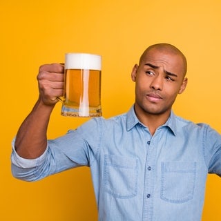 Ein Mann schaut skeptisch auf ein Glas Bier und eine Flasche Bier in seiner Hand. Ein alkoholfreies Getränk kann auch Alkohol enthalten.